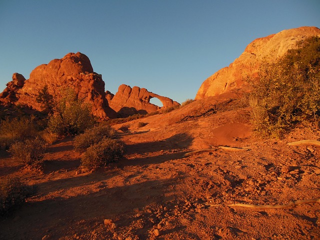 Sandstone arch on a distant ridge glowing orange in late light, with red rock and desert scrub in the foreground