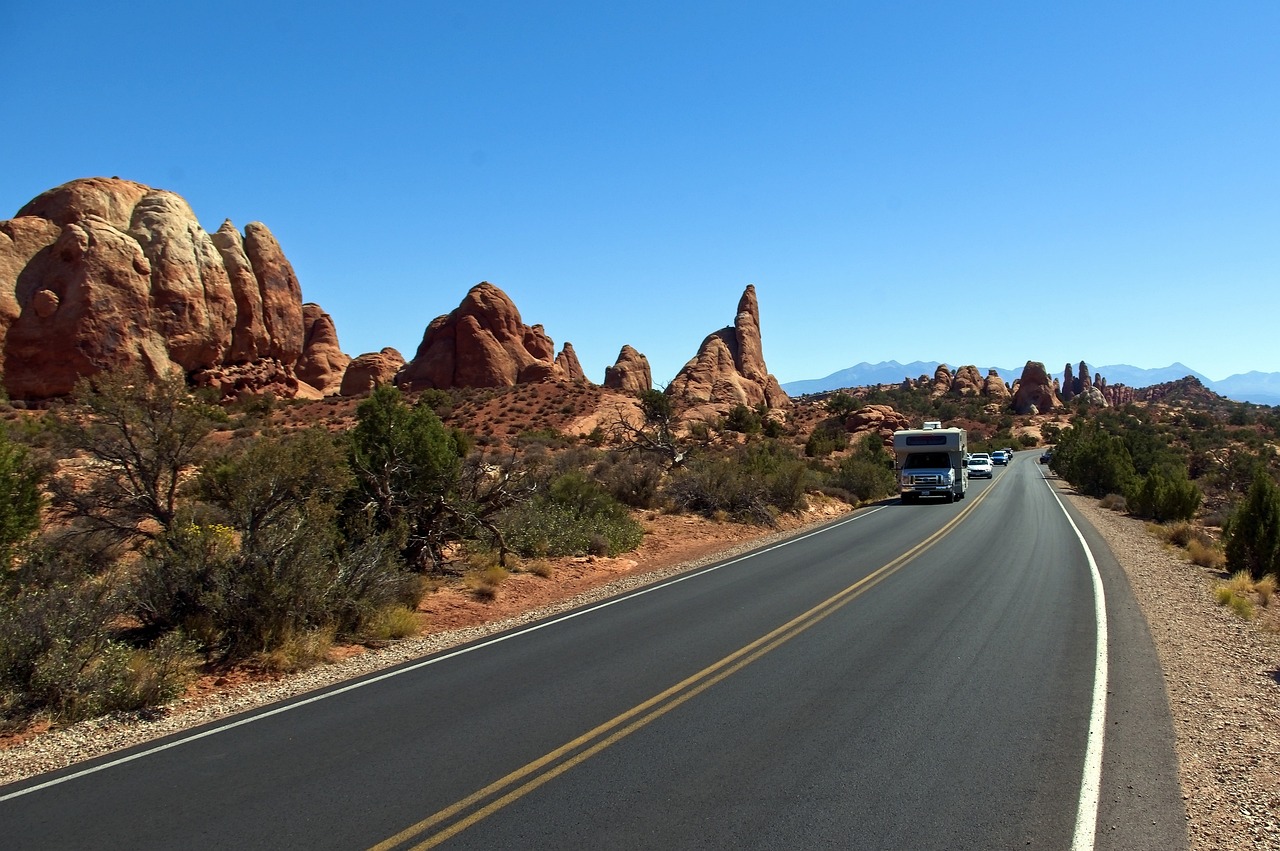Paved park road with a white RV and cars heading toward red rock spires and distant mountains under a clear blue sky