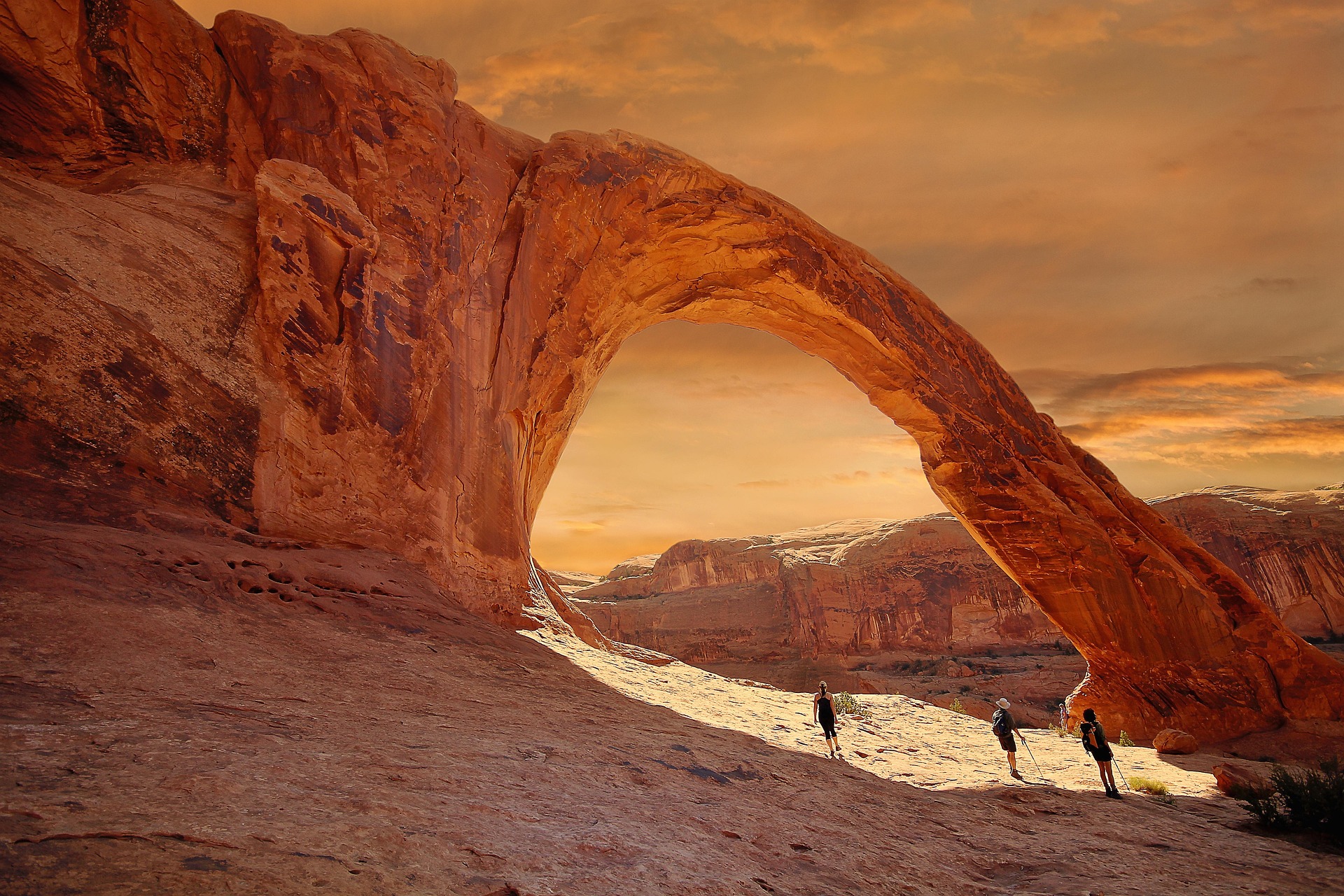 Three hikers walk beneath Corona Arch under an orange sunset sky