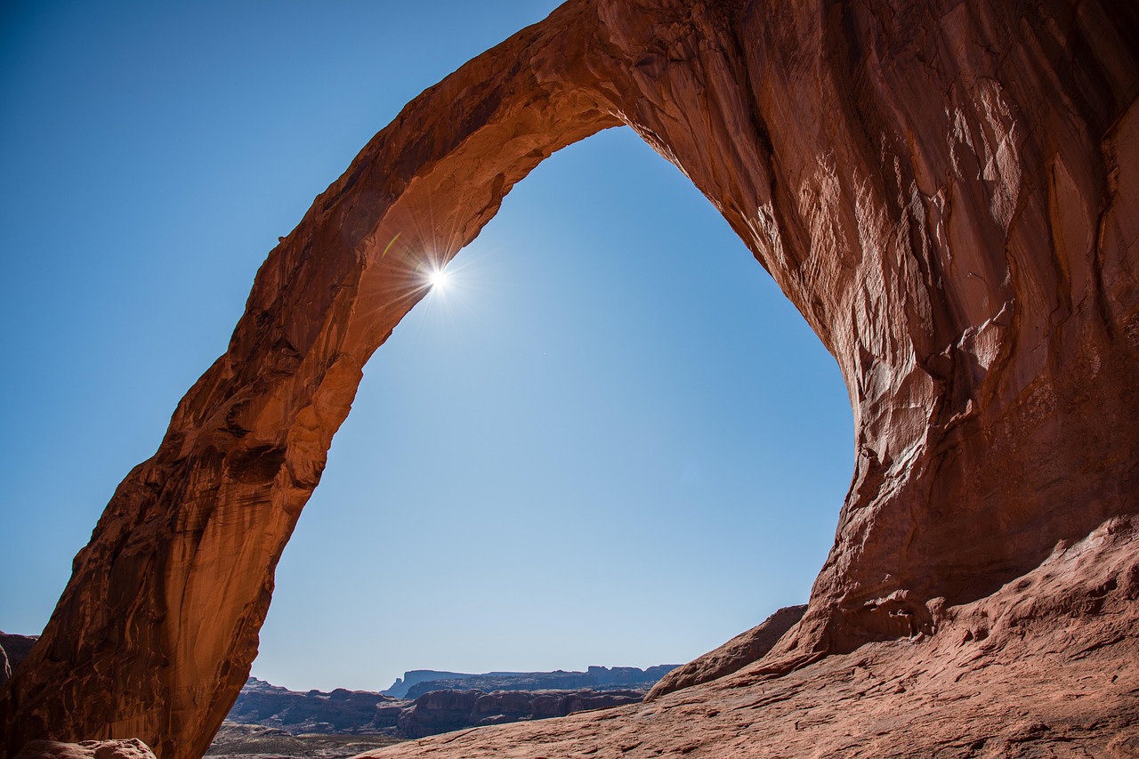 Corona Arch curving overhead with the sun as a starburst against a clear blue sky and a distant canyon rim