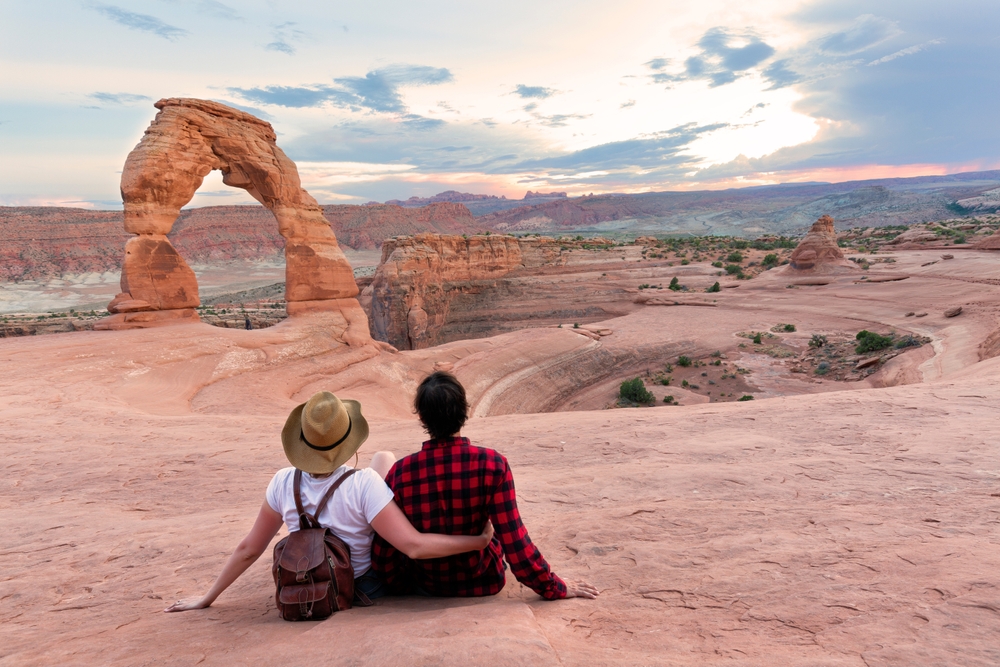 Two hikers sit together on slickrock looking at Delicate Arch at dusk