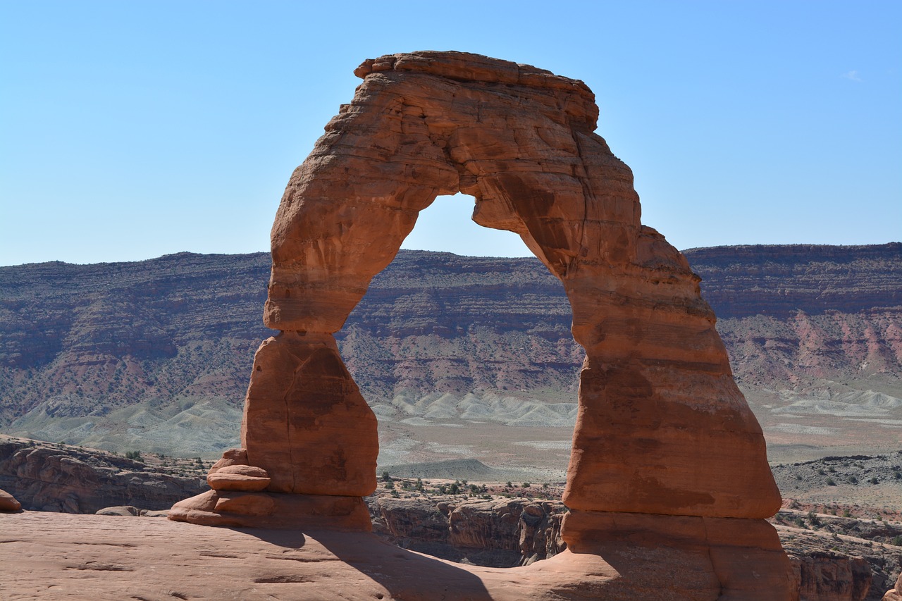 Delicate Arch against a clear blue sky with a layered canyon wall and pale desert basin behind it