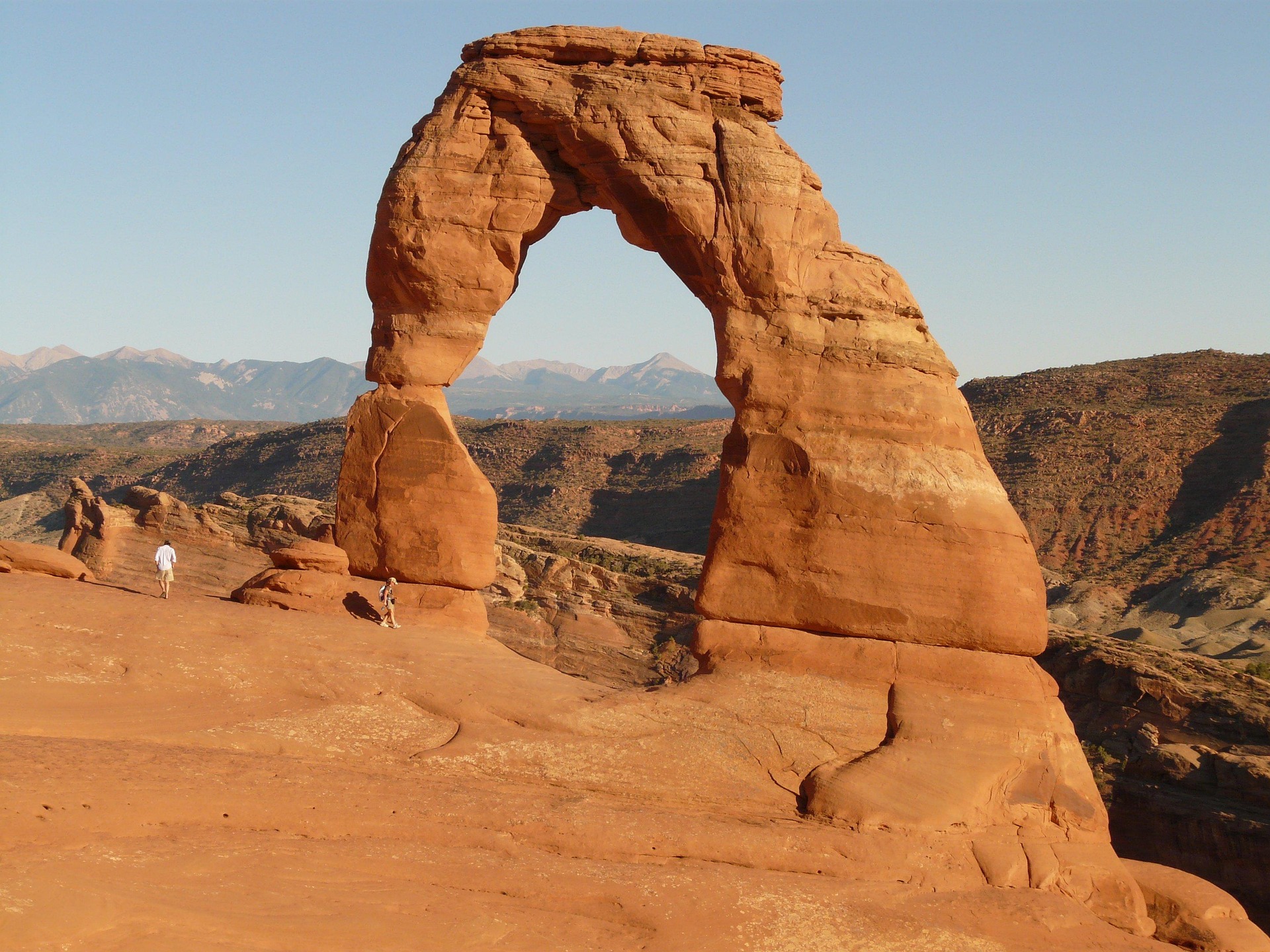Delicate Arch in warm light with two visitors on the slickrock and the La Sal Mountains behind