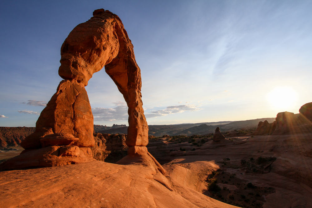 Delicate Arch standing on slickrock with the low sun setting beside it