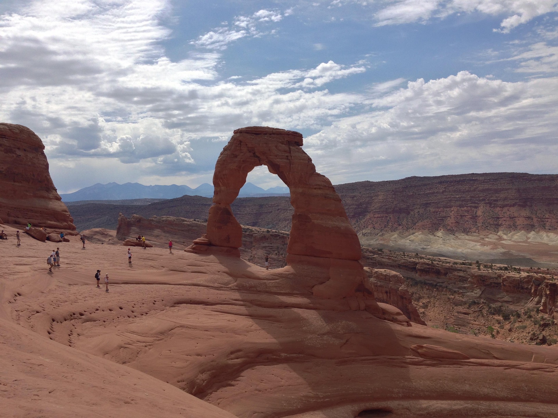 Delicate Arch with visitors on the slickrock bowl and the La Sal Mountains behind under scattered clouds