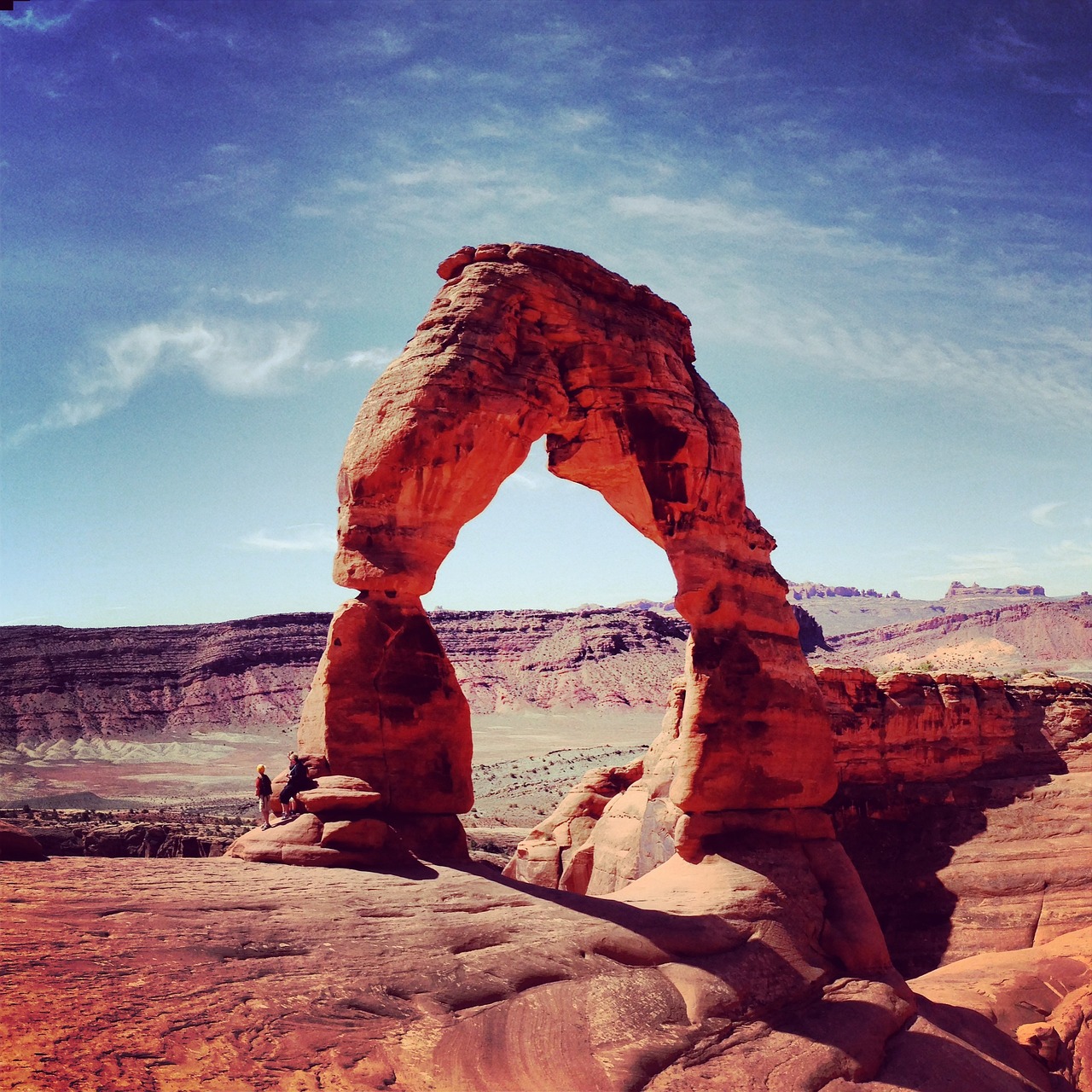 Delicate Arch glowing red under a deep blue sky with two small visitors sitting at its base on the left
