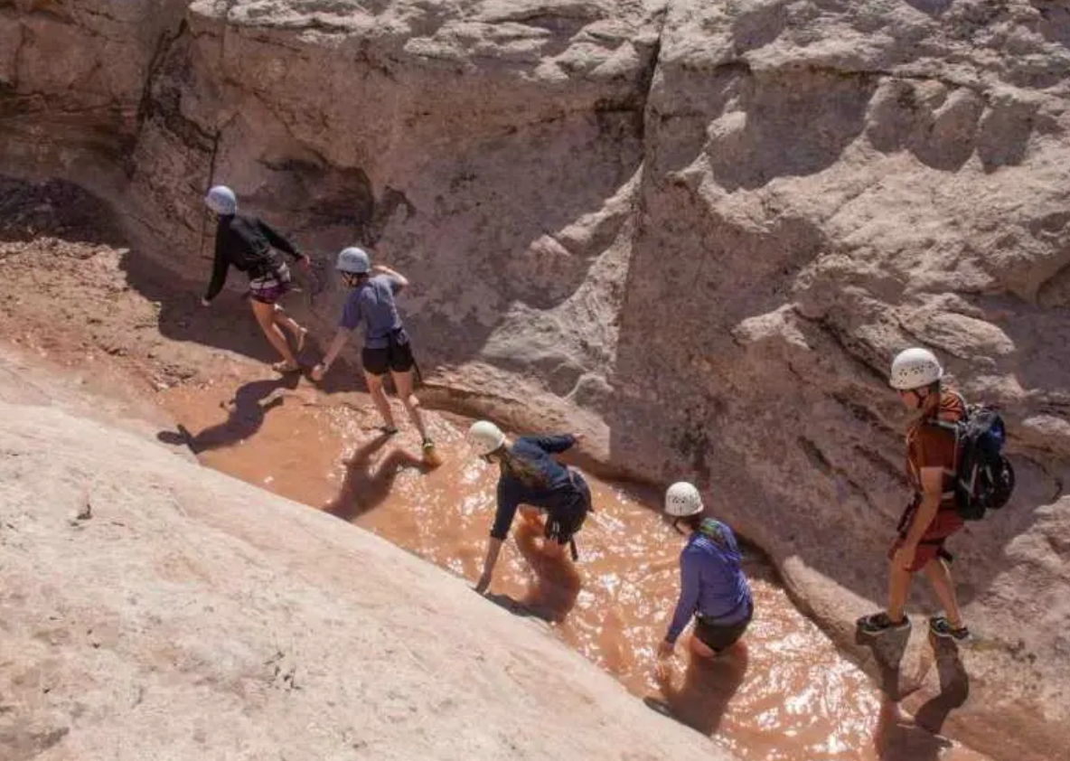 Five people in helmets wading and scrambling through a muddy pool in a pale sandstone slot canyon