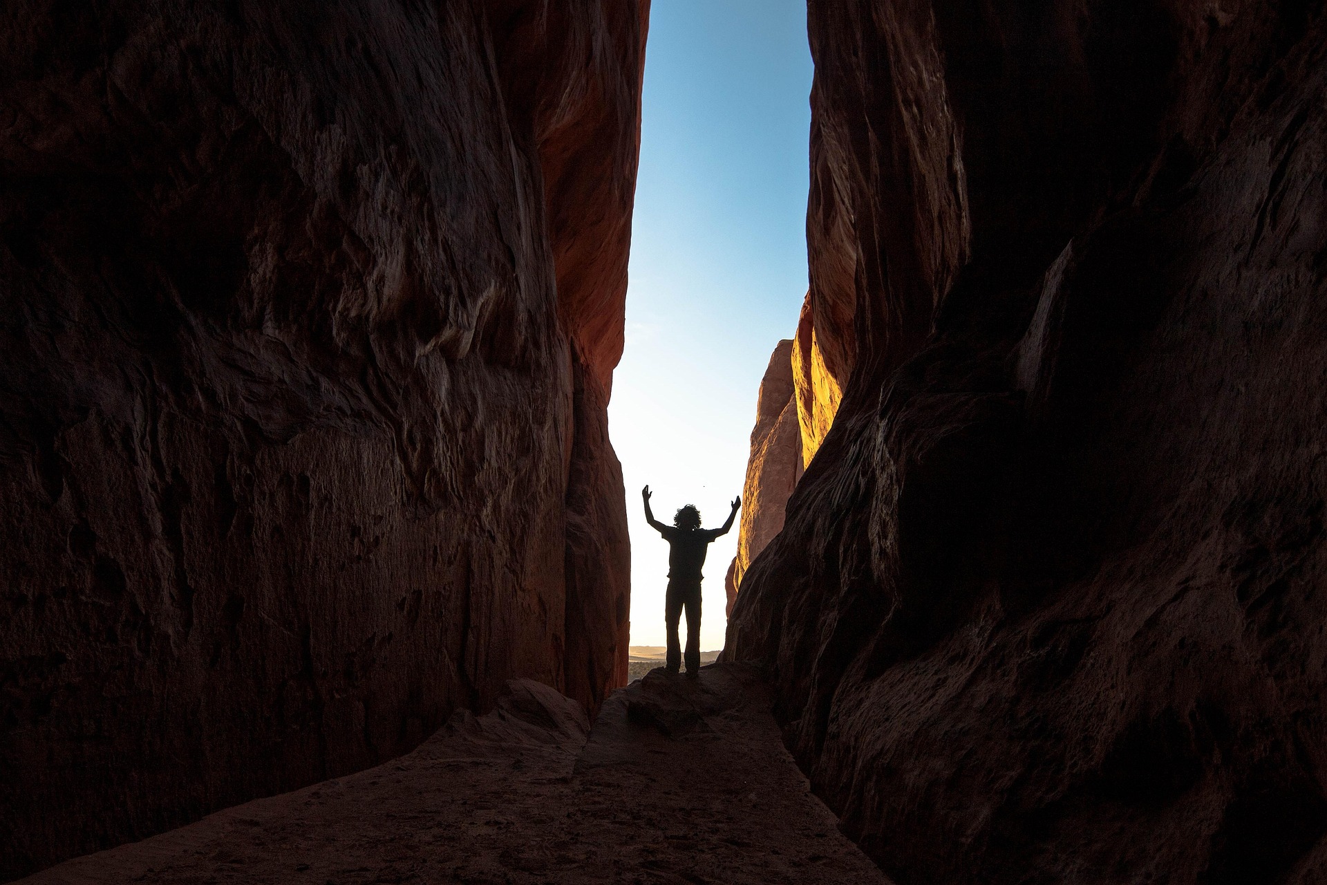 Silhouetted hiker with arms raised in a narrow passage between tall dark red sandstone walls