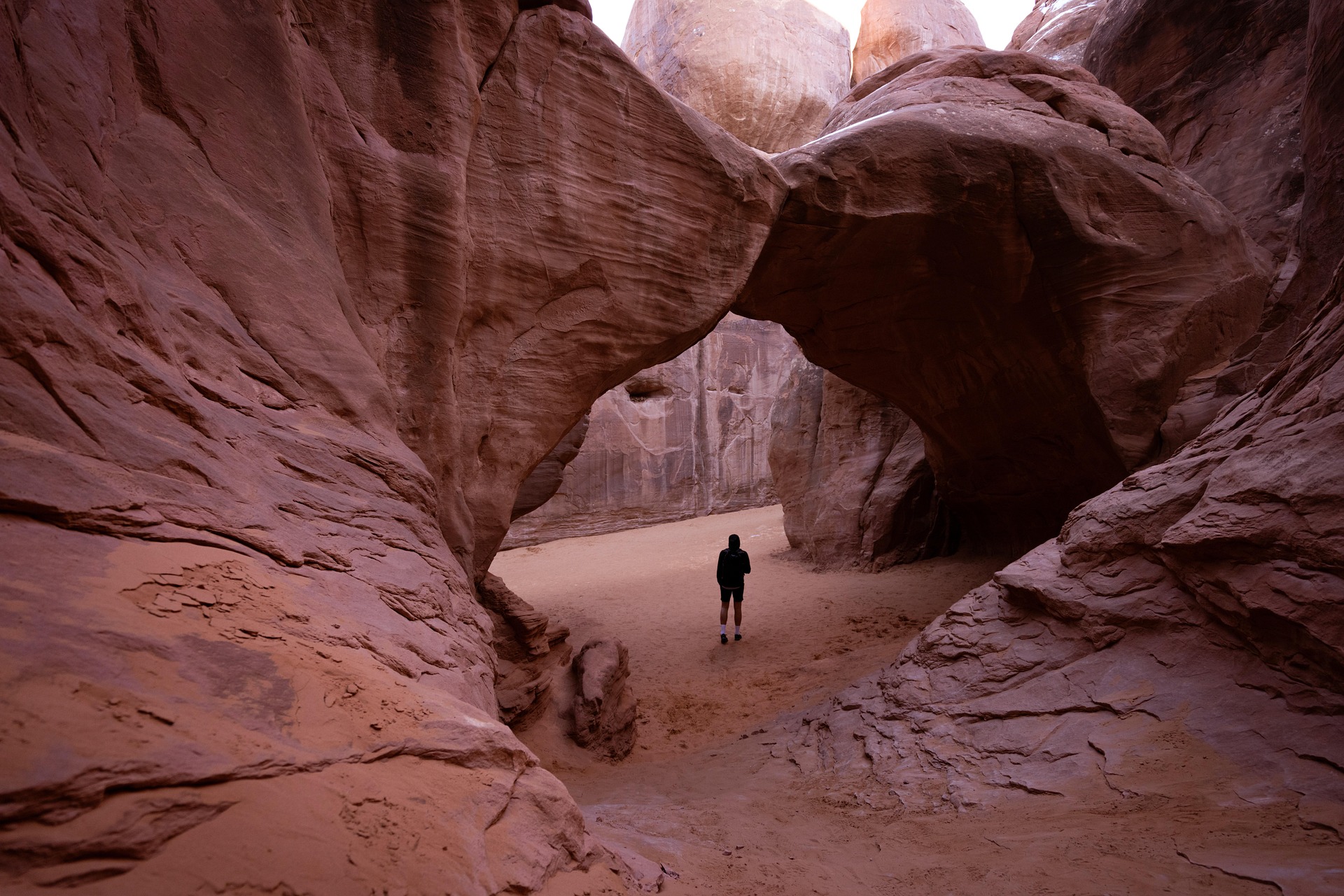 Hiker standing on sand beneath a large sandstone arch between tall red rock walls