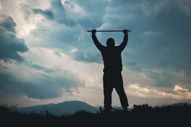 Hiker silhouetted against a stormy sky raising a trekking pole overhead on a grassy ridge