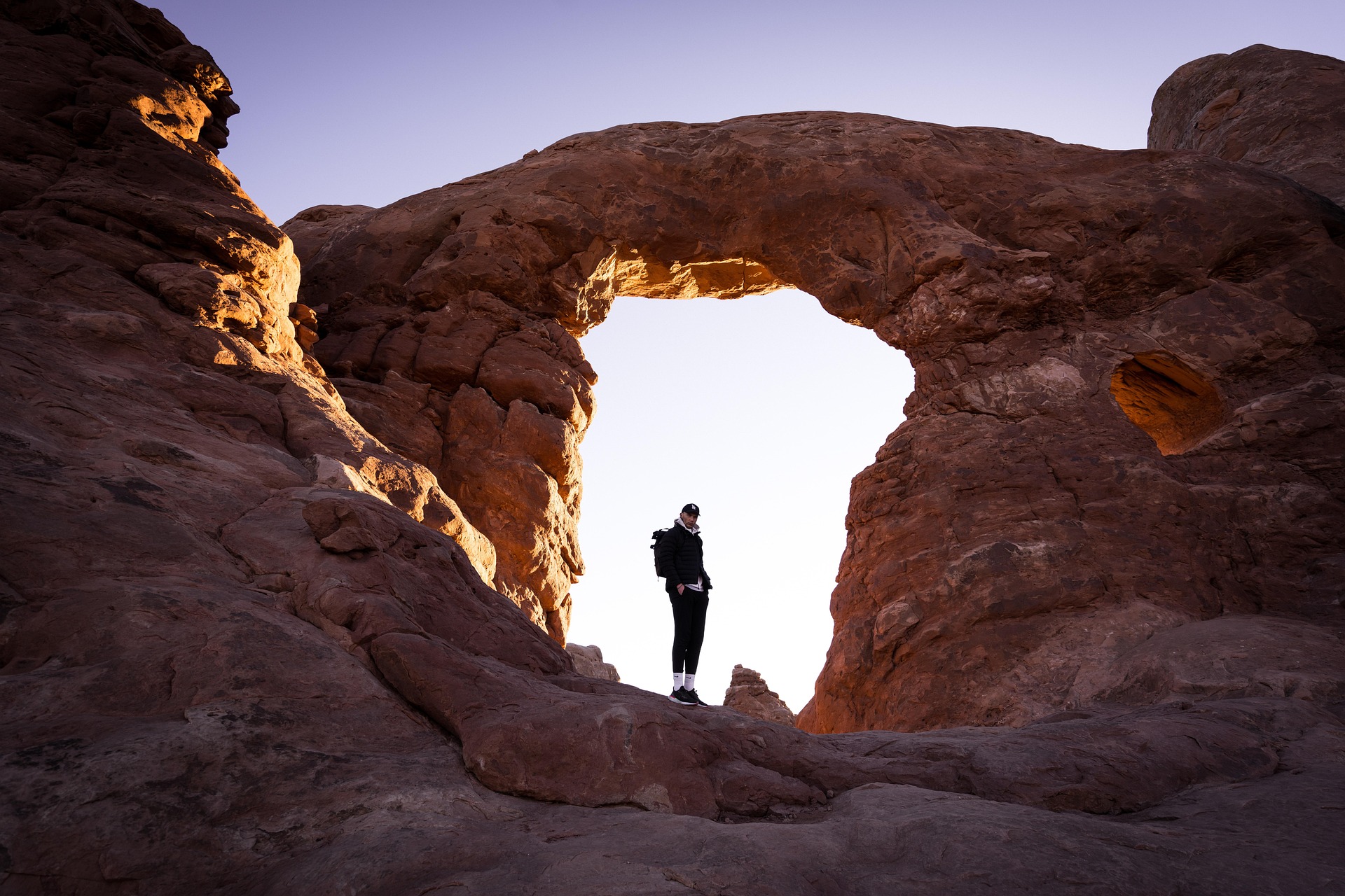 Hiker with a backpack standing in the opening of a sandstone arch at twilight