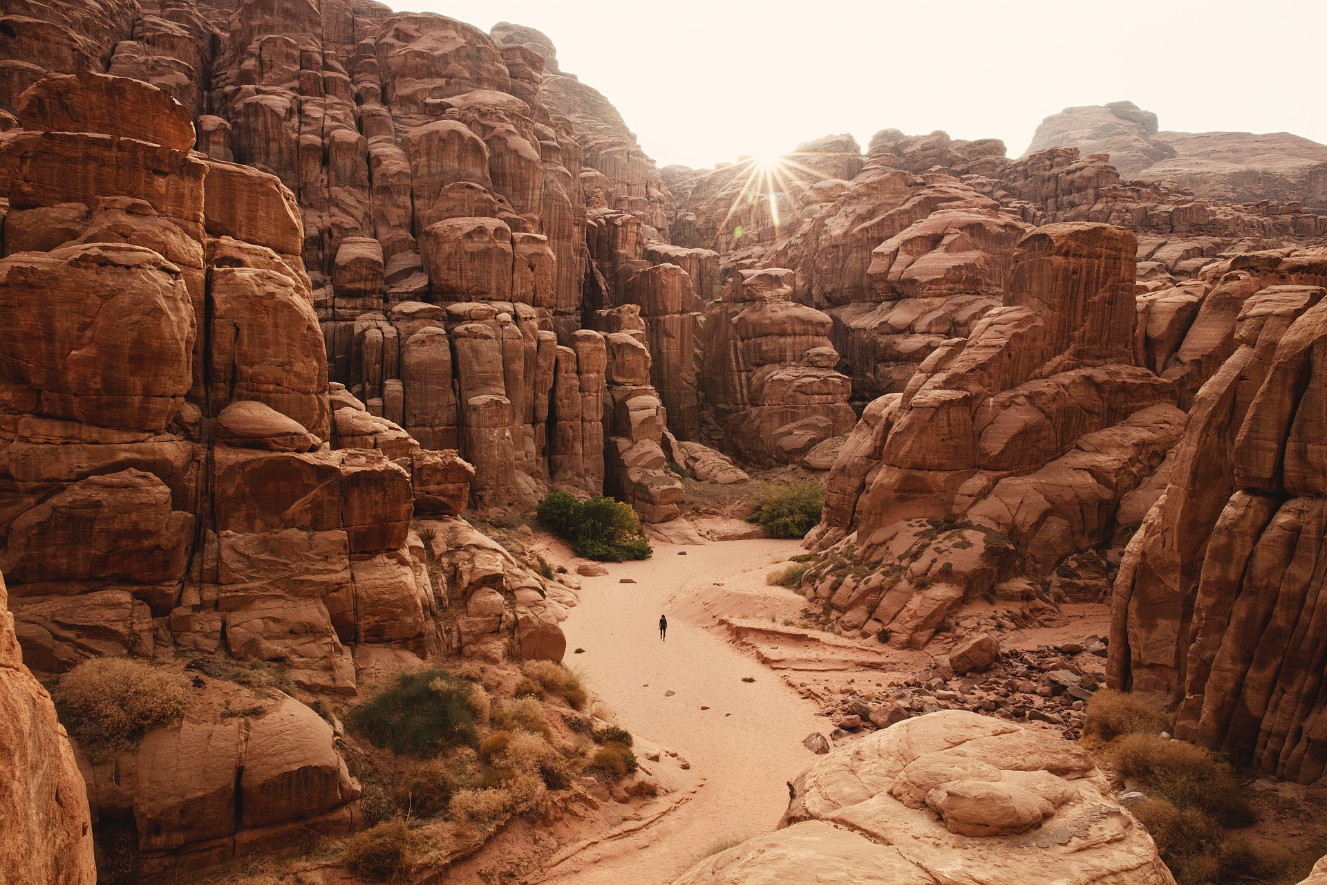 Lone hiker walking a sandy wash between tall layered sandstone cliffs with the sun starburst above