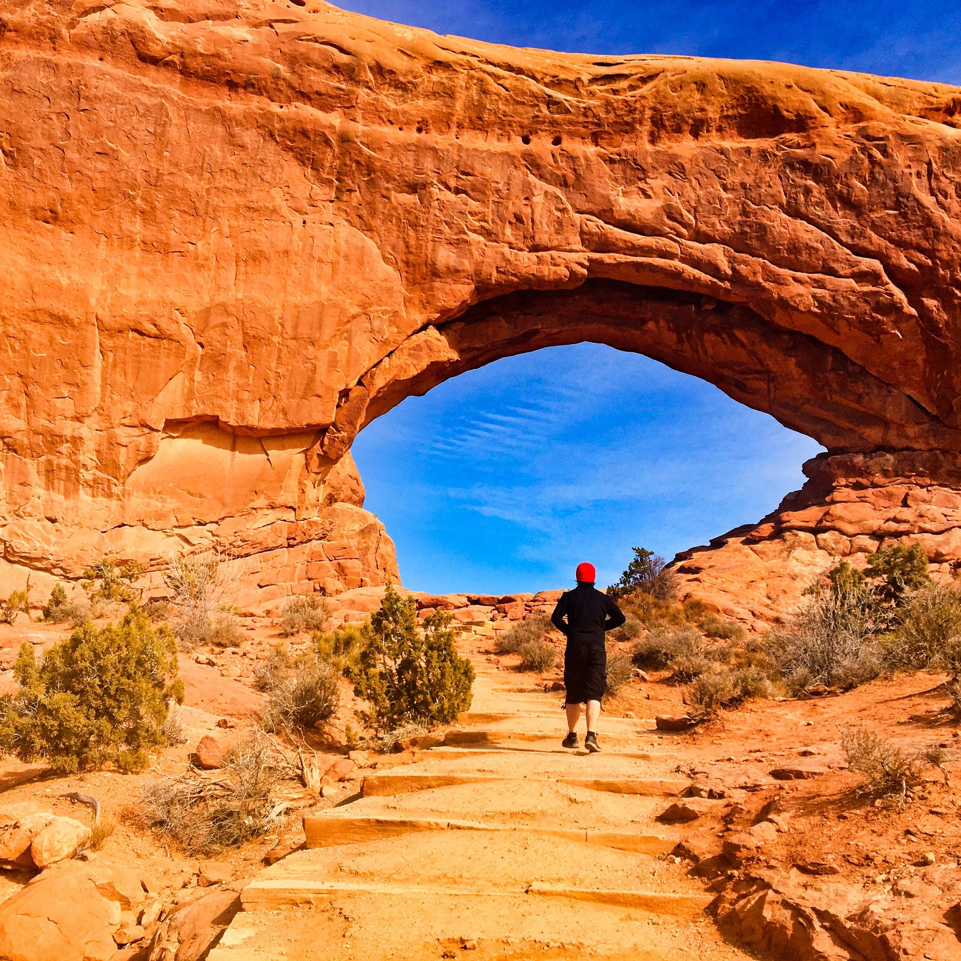 Hiker in a red hat walking up a stepped sandstone trail toward a large arch with blue sky in the opening