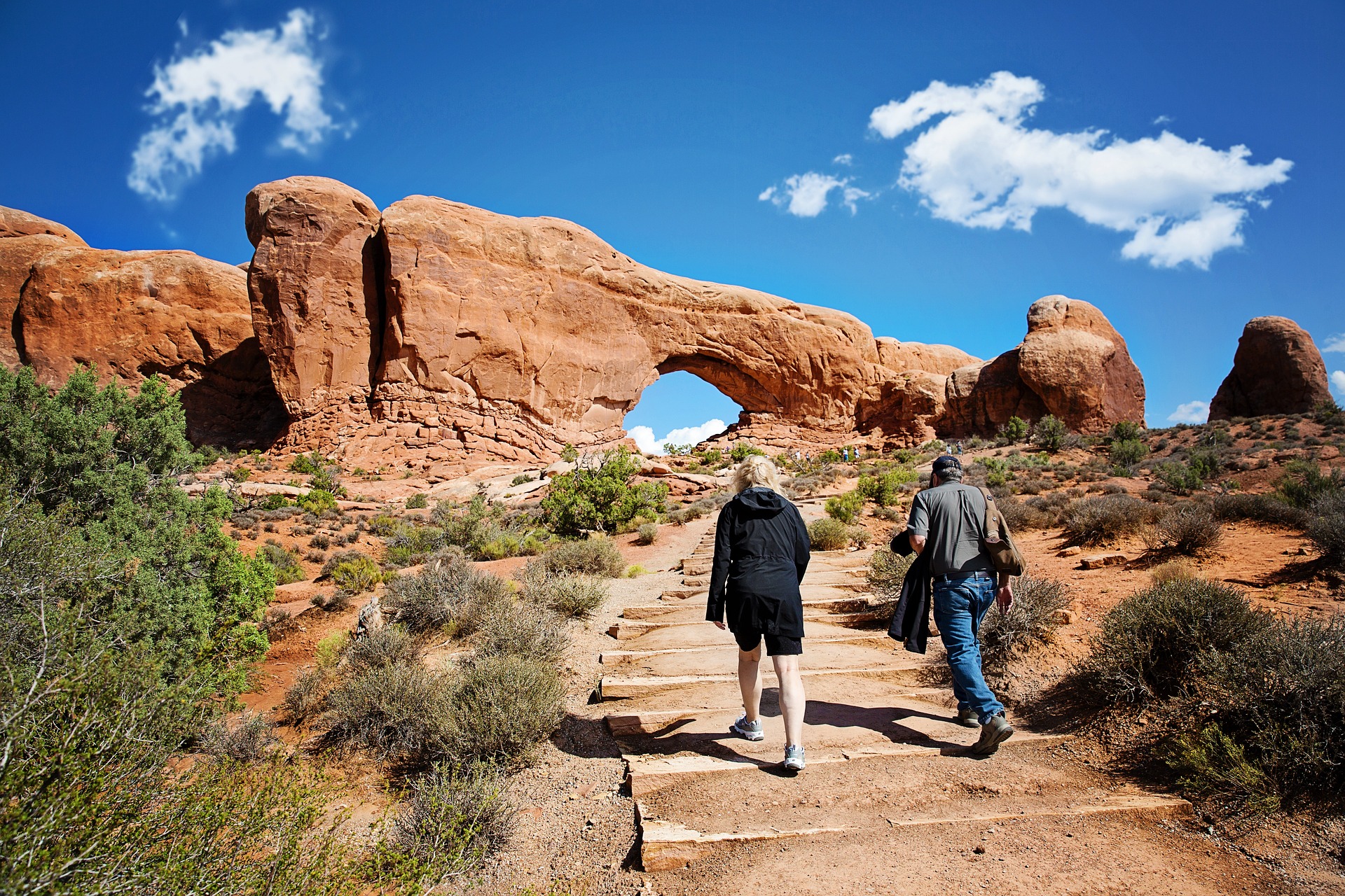 Two hikers climb a stone stepped trail toward North Window arch under a bright blue sky and white clouds