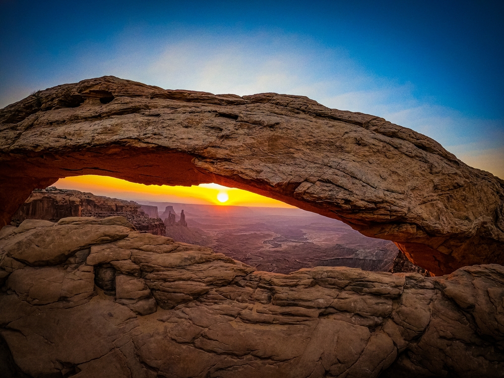 Sun rising through the opening of Mesa Arch with distant canyon spires glowing orange