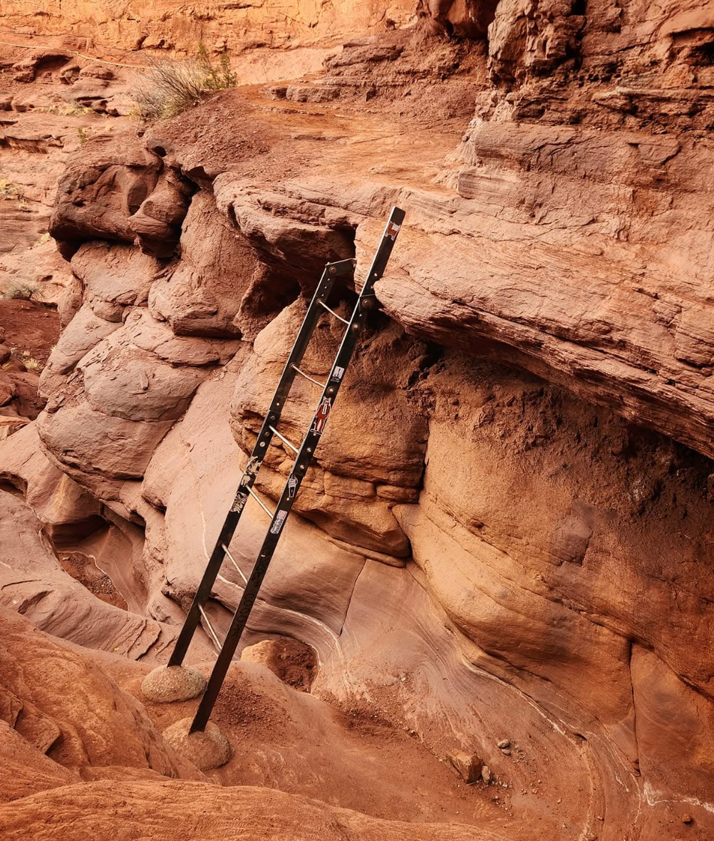 Steel ladder with stickers leaning against a red sandstone ledge above a narrow, curved slot