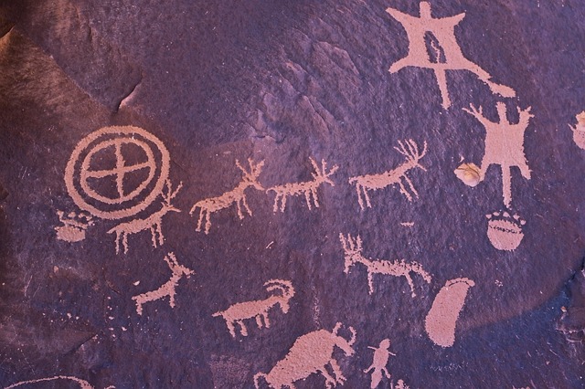 Ancient petroglyphs on dark desert varnish, including a circle, horned animals, a bighorn sheep and a human figure