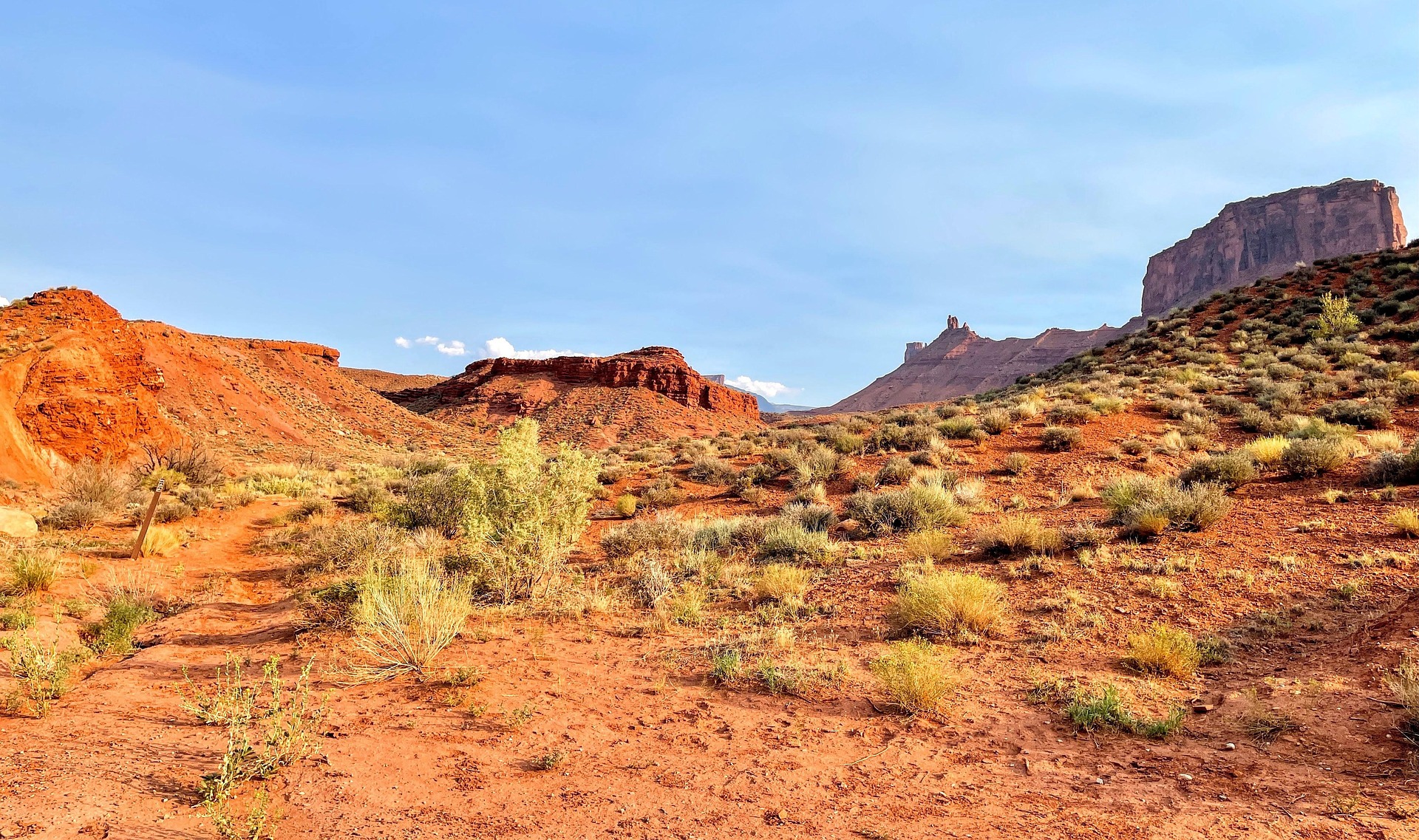 Red Desert Trail Below the Buttes Red dirt desert with sagebrush, a trail marker post at left and sandstone buttes under a pale blue sky