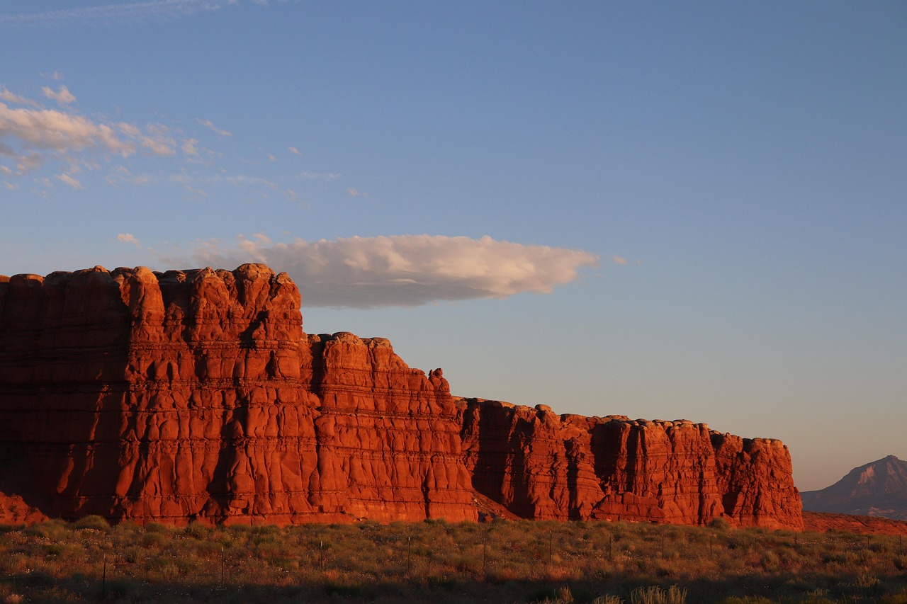 Red Cliff Wall at Sunset Layered red sandstone cliff wall glowing in low sun with a single cloud overhead and a distant peak at right