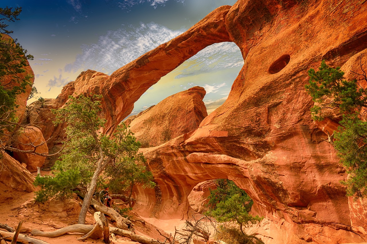 Sandstone arch with two spans and a small round window, framed by juniper trees and dead wood under a vivid sky